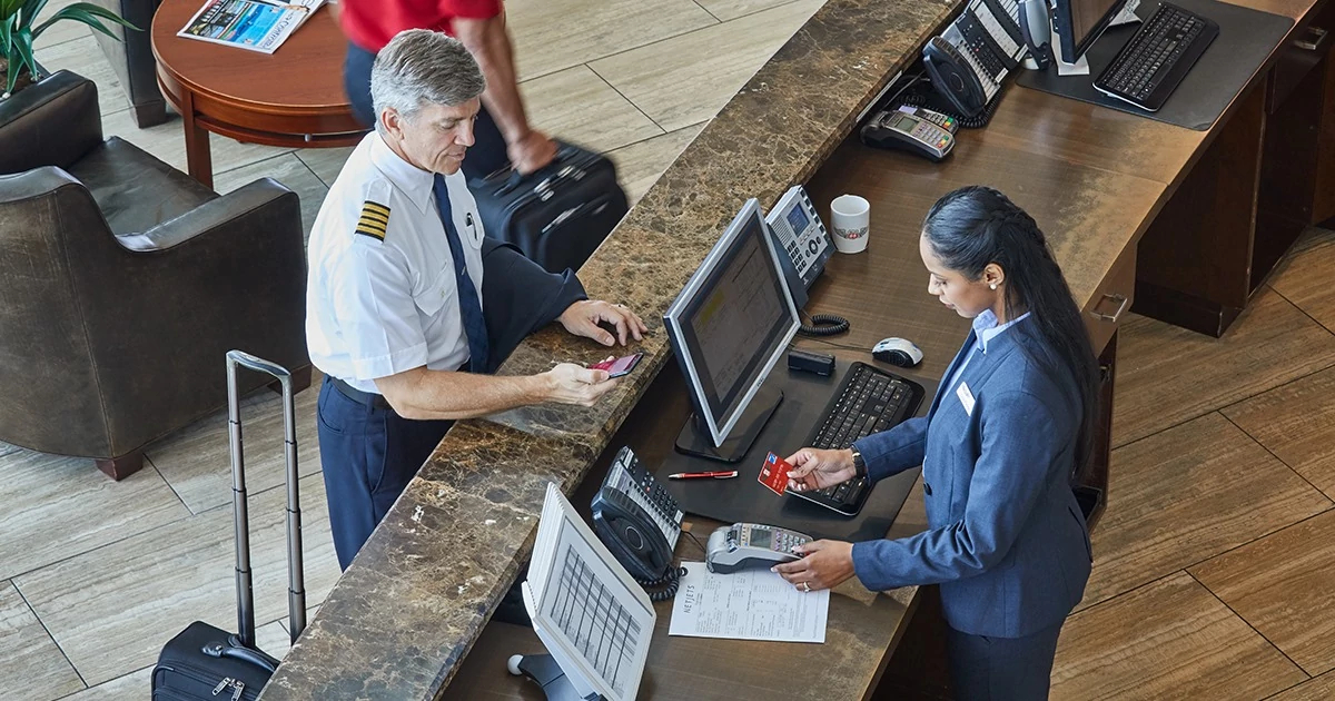 A pilot in uniform presenting a credit card at a front desk to a female attendant, highlighting credit card programs for pilots and businesses at an FBO.